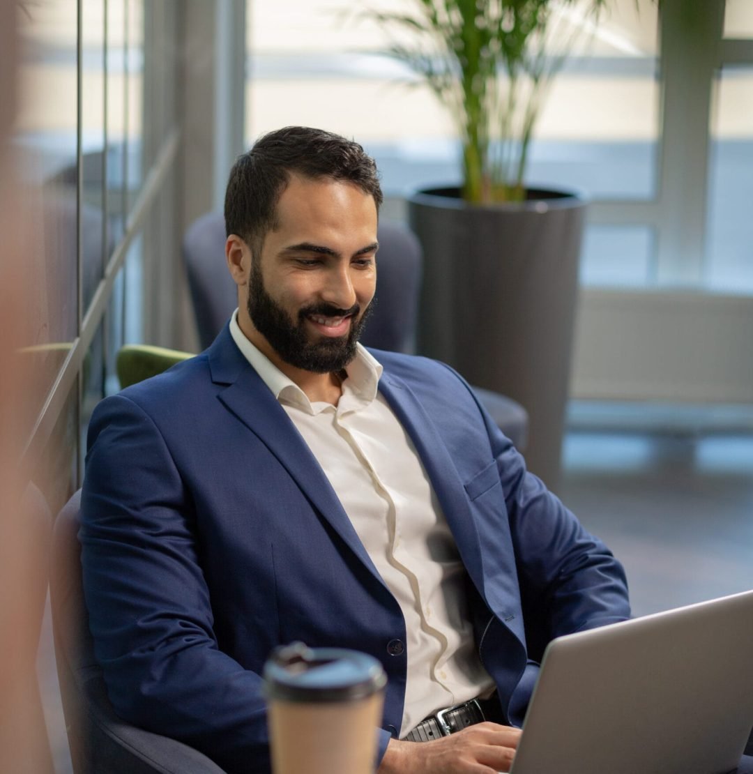 Handsome brunette male person having video conference Cheerful office worker keeping smile on his face while working with his laptop